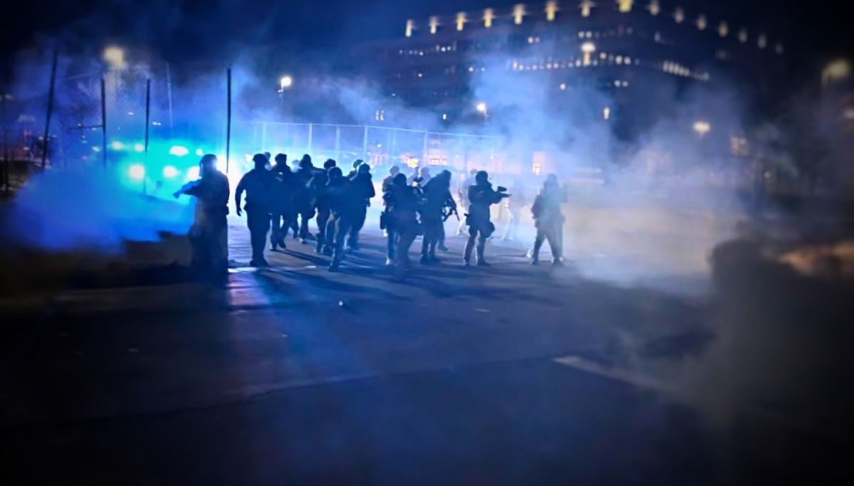 A line of officers advances through drifting gas lit blue by emergency lights at night.
