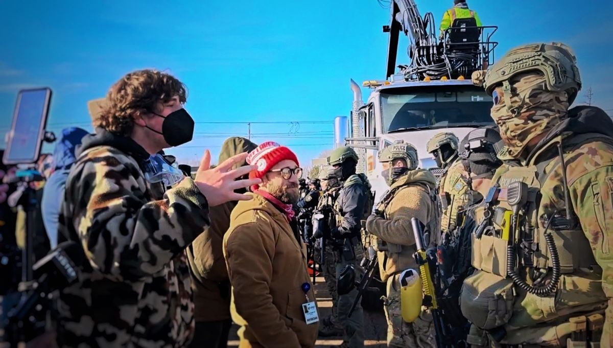 A masked demonstrator gestures while speaking to a line of soldiers in front of an armoured truck under a clear sky.