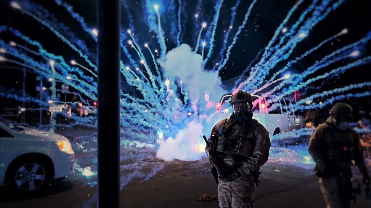 Fireworks burst in an arc across a street as an officer in a gas mask walks toward the camera at night.