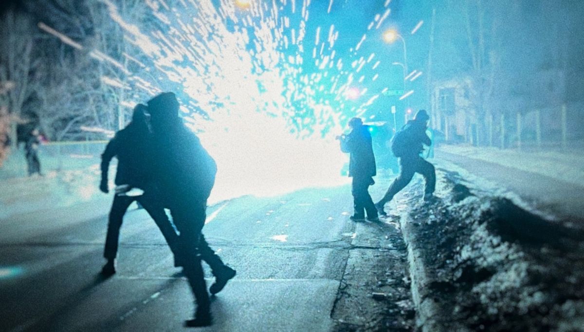 Silhouetted figures scatter on a snow-lined street as a bright flash detonates behind them.