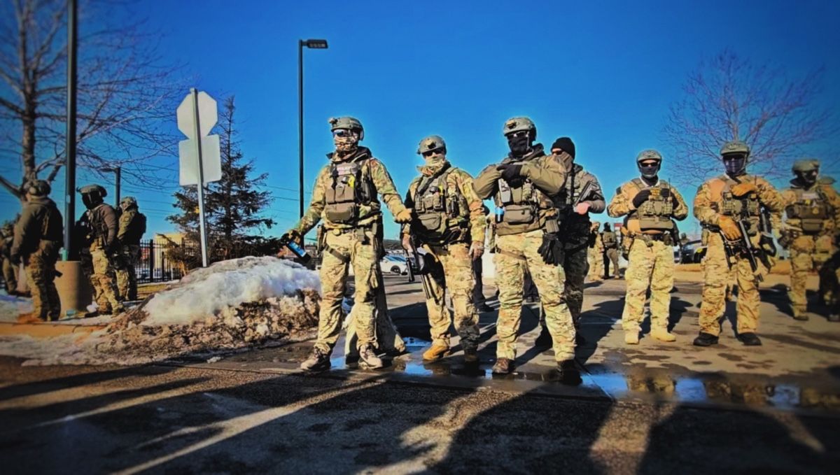 Soldiers in camouflage and face coverings stand in a loose line on a sunlit street beside snow banks.